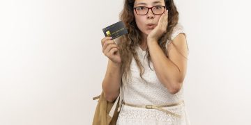jovem bonita impressionada usando oculos e bolsa traseira segurando um cartao de credito com mao no rosto isolado no branco